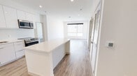 This single-floor residence showcases a clean, modern design with a bright, open-concept layout and cohesive contemporary finishes throughout. The heart of the space is the kitchen, which features a large central island with a white quartz countertop, high-gloss white cabinetry extending to the ceiling, a stone tile backsplash, and a full suite of stainless steel appliances including a refrigerator, range, dishwasher, and built-in microwave. The kitchen flows seamlessly into the open living area, which is bathed in natural light from large windows and finished with warm wood-look flooring and recessed lighting overhead. Two well-proportioned bedrooms each offer wide windows that bring in abundant natural light, wood-look flooring, ceiling fans, and sliding closet doors for ample storage. The two bathrooms are finished in a consistent modern palette, featuring white floating vanities with quartz countertops, backlit mirrors, tile flooring, and accessible grab bars. One bathroom includes a double-sink vanity and a walk-in tiled shower. Throughout the residence, crisp white walls, clean lines, and quality materials come together to create a bright, airy, and functional living environment.