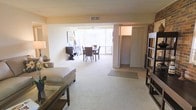 This single-floor residence offers an open, flowing layout that seamlessly connects the living and dining areas into one cohesive space. The living area is finished with soft, plush wall-to-wall carpeting that adds warmth underfoot, while the open floor plan creates a sense of generous spaciousness throughout. Large sliding glass doors at the far end of the room flood the interior with abundant natural light and provide views of the greenery outside, enhancing the bright and airy ambiance. A ceiling fan is positioned above the dining area, offering both comfort and a casual, functional touch. The dining space comfortably accommodates a full table and chair set, making it well-suited for everyday meals and entertaining. A wide closet with bifold doors is conveniently situated adjacent to the main living space, offering ample storage. An exposed brick accent wall adds a touch of character and texture to the otherwise clean, neutral interior palette. The walls are finished in a soft, warm tone that complements the natural light throughout. Overall, the space presents a practical and comfortable single-level layout with a welcoming atmosphere.
