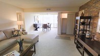 This single-floor residence offers an open, flowing layout that seamlessly connects the living and dining areas into one cohesive space. The living area is finished with soft, plush wall-to-wall carpeting that adds warmth underfoot, while the open floor plan creates a sense of generous spaciousness throughout. Large sliding glass doors at the far end of the room flood the interior with abundant natural light and provide views of the greenery outside, enhancing the bright and airy ambiance. A ceiling fan is positioned above the dining area, offering both comfort and a casual, functional touch. The dining space comfortably accommodates a full table and chair set, making it well-suited for everyday meals and entertaining. A wide closet with bifold doors is conveniently situated adjacent to the main living space, offering ample storage. An exposed brick accent wall adds a touch of character and texture to the otherwise clean, neutral interior palette. The walls are finished in a soft, warm tone that complements the natural light throughout. Overall, the space presents a practical and comfortable single-level layout with a welcoming atmosphere.