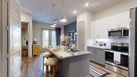 This thoughtfully designed one-bedroom, one-bathroom residence offers an efficient and stylish layout across a single floor. The heart of the home is the open-concept kitchen, which features two-tone cabinetry — crisp white uppers paired with gray lowers — stainless steel appliances, a subway tile backsplash, and a generous granite-topped island with an integrated sink and seating for two. Pendant lighting illuminates the island, creating a warm and functional gathering space. The kitchen flows seamlessly into the living area, which is accented by a bold slate-blue feature wall, hardwood-style flooring, and a sliding glass door that invites natural light from outside. The bedroom is a serene retreat highlighted by a dark wood-paneled accent wall, warm globe wall sconces, and a window with natural light. It connects directly to a dedicated closet with built-in hanging rods for organized storage. The full bathroom is well-appointed with a granite-topped gray vanity, a full soaking tub with tiled surround, and hardwood-style flooring that ties together the cohesive design aesthetic found throughout the residence.