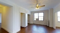 This single-floor space showcases a blend of classic architectural character and practical design. The main living area is defined by rich, dark hardwood floors that run throughout, lending warmth and timeless appeal to the interior. Crown molding and decorative wall paneling add refined detailing to the white-painted walls, creating a clean yet elegant aesthetic.

A ceiling fan with a built-in light fixture serves as a functional centerpiece overhead, providing both air circulation and ambient lighting. Natural light enters through a double-pane window fitted with an air conditioning unit, offering both ventilation and climate control while framing views of the surrounding greenery outside.

Storage is notably well-considered, with two closets side by side — one featuring open built-in shelving, ideal for organized storage, and the other a standard enclosed closet. The closets are framed by paneled white doors that complement the overall architectural style of the space.

The layout is open and uncluttered, allowing for flexible furniture arrangements. The combination of classic detailing, functional storage solutions, hardwood flooring, and natural light makes this a well-appointed and inviting space suitable for a variety of uses.