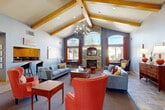 Located at 3775 W 25th St in Greeley, Colorado, this single-floor community clubhouse is a well-appointed gathering space defined by its dramatic architectural character and warm, inviting atmosphere. The centerpiece of the main lounge is a soaring vaulted ceiling adorned with bold, honey-toned exposed wood beams, beneath which hangs an elegant chandelier that anchors the room with a touch of refined style. A floor-to-ceiling stacked stone fireplace with an arched surround serves as a striking focal point, framed by tall arched windows dressed in rich burnt-orange curtains that flood the space with natural light. The open layout accommodates multiple seating arrangements, featuring plush gray sofas, vibrant coral accent chairs, and wooden coffee tables, creating distinct yet cohesive conversation areas. The flooring transitions seamlessly between slate tile and plush carpet, adding texture and warmth throughout. A kitchenette with warm wood cabinetry is integrated into the space, offering added functionality for events and gatherings. Colorful original artwork and decorative mirrors punctuate the cool blue-gray walls, contributing to the space's energetic yet comfortable ambiance. Large windows and glass entry doors frame outdoor views, ensuring the interior remains bright and connected to its surroundings.