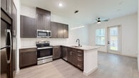 This single-floor unit showcases a contemporary design with a cohesive blend of dark cabinetry, light wood-look flooring, and crisp white walls throughout the main living areas. The kitchen serves as a central highlight, featuring dark espresso-toned shaker cabinets, a white subway tile backsplash, stainless steel appliances including a range, microwave, and dishwasher, and a peninsula with granite-style countertops and an undermount sink with a matte black faucet. A linear pendant light fixture illuminates the peninsula, complementing the recessed lighting above. The kitchen opens directly into the living area, which benefits from natural light through a window and a glass-paneled exterior door providing access to an outdoor space. A ceiling fan is centrally positioned in this area.

The bedrooms are finished with plush carpet and ceiling fans, with generous windows that bring in ample natural light. The primary bathroom is well-appointed with matching dark cabinetry, a granite-topped vanity, a tall linen tower, and an adjacent walk-in closet. The bathroom also features light wood-look flooring, black accent hardware, and a separate shower and toilet area, offering a clean, functional layout throughout.