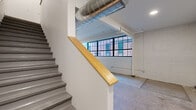 Located at 601 W 2nd St in downtown Davenport, Iowa, this two-floor urban loft blends industrial character with modern finishes. The main level features polished terrazzo flooring, exposed structural beams, and prominent large-pane industrial windows that frame views of the surrounding brick buildings, filling the space with natural light. Exposed cylindrical ductwork runs overhead, lending an authentic loft aesthetic to the open layout. The well-appointed kitchen showcases dark wood cabinetry, granite-style countertops, and a full suite of stainless steel appliances including a range, microwave, dishwasher, and refrigerator, all complemented by painted brick accent walls and terrazzo floors. A staircase with a warm wood handrail connects the two levels. The upper floor offers carpeted flooring, a ceiling fan, and painted brick walls, creating a quieter, more intimate atmosphere. A dedicated bathroom on the upper level includes a dark wood vanity with granite countertops, a walk-in shower, and light wood-look flooring. A closet provides convenient storage. The combination of industrial heritage details and contemporary updates gives this Davenport loft a distinctive character well-suited to urban living.