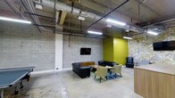 This single-floor space embraces an industrial aesthetic, blending raw architectural elements with thoughtfully curated furnishings to create a unique and functional environment. The exposed ceiling reveals an intricate network of ductwork, conduit, and structural beams, lending the space an open, loft-like character. Polished concrete floors run throughout, reinforcing the industrial feel while providing a durable, low-maintenance surface.

The lounge area serves as the focal point of the space, anchored by a tufted black leather sofa and complementary seating including cushioned accent chairs gathered around a wooden coffee table. A patterned area rug defines the seating zone and adds warmth to the otherwise raw surroundings. A wall-mounted flat-screen television is positioned on the concrete masonry block wall, making the area suitable for both casual gatherings and presentations.

Bold design choices inject personality into the space — a vivid chartreuse accent wall provides a striking contrast against the neutral grey of the exposed block walls, while a decorative botanical-patterned wallcovering adds texture and visual interest to an adjacent corner. Overhead fluorescent strip lighting ensures the space is well-illuminated throughout. Overall, this space strikes a compelling balance between utilitarian industrial design and inviting, stylish comfort.