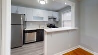 Located at 122 Maybrook Dr in Maywood, NJ, this thoughtfully updated single-floor unit offers a clean, modern aesthetic with neutral gray tones and quality finishes throughout. The kitchen is a standout feature, boasting white shaker-style cabinetry, a classic subway tile backsplash, light quartz countertops, and a full suite of stainless steel appliances including a gas range, dishwasher, and refrigerator. A peninsula counter with seating overhang provides additional workspace and casual dining options, while light hardwood-style flooring anchors the space. The carpeted bedrooms are generously proportioned, each painted in soothing gray hues and equipped with natural light from windows fitted with horizontal blinds. Recessed lighting illuminates the larger bedroom, which also features a staircase landing area, while a classic flush-mount fixture lights the second. Radiator heating is present in both bedrooms, ensuring year-round comfort. The bathroom has been fully renovated with large-format white wall tiles, a soaking tub with shower, a dark-cabinetry vanity with a round mirror, and crisp white tile flooring. The overall layout, as seen in the dollhouse view, is functional and well-organized, making this a polished and move-in-ready space.