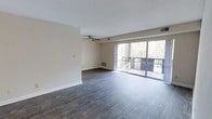 This single-floor space offers a clean, modern aesthetic characterized by an open and airy layout. The main living area is finished with rich, dark hardwood-style flooring that provides a striking contrast against the neutral, light-toned walls and white baseboards, creating a polished and contemporary feel throughout.

A ceiling fan is centrally positioned overhead, contributing both functional airflow and a subtle design element to the living space. Natural light floods the interior through a large sliding glass door that spans a generous portion of the exterior wall, offering unobstructed views of the lush, tree-lined surroundings beyond the private balcony. The balcony, enclosed by a metal railing, extends the living experience outdoors and provides a serene setting with views of mature trees.

The space also features wall-mounted vents for climate control, ensuring year-round comfort. The open floor plan allows for flexible furniture arrangement, making it well-suited for a variety of lifestyle needs. The overall ambiance is bright, calm, and inviting, with the combination of natural light, neutral tones, and quality flooring lending the space a refreshed and move-in ready quality.