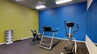 This single-floor fitness space is designed to support cardiovascular exercise in a focused, energetic environment. The room features a bold two-tone color scheme, with a vivid blue accent wall meeting a bright yellow-green wall, creating a visually stimulating atmosphere that complements an active workout setting. The flooring consists of durable rubber gym matting, providing cushioning and traction suited to exercise equipment use. The space is equipped with essential cardio machines, including a treadmill, a stationary spin bike, and an elliptical trainer, offering a variety of low- and high-impact workout options. Overhead, a ceiling fan ensures air circulation throughout the room, accompanied by fluorescent lighting that keeps the space well-lit. The layout is functional and straightforward, allowing users easy access to each piece of equipment. The clean, uncluttered design keeps the focus firmly on fitness, making this gym room a practical and purpose-driven facility for regular physical activity.