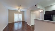 This single-floor space showcases an open-concept layout that seamlessly connects the living area and kitchen. The living room is finished with rich, dark hardwood-style flooring that extends throughout, creating a warm and cohesive aesthetic. Neutral gray walls provide a clean, modern backdrop, while a ceiling fan with integrated lighting ensures year-round comfort. French doors along the far wall invite natural light into the space and offer direct access to an outdoor area, enhancing the sense of openness.

The kitchen is partially visible from the living area, separated by a raised bar counter finished with granite-style countertops, adding a touch of elegance and providing a casual dining or prep surface. The kitchen features its own dedicated lighting, maintaining a bright and functional workspace.

A louvered bi-fold door indicates convenient in-unit storage or a closet, contributing to the overall practicality of the layout. HVAC vents are integrated into the ceiling throughout, supporting efficient climate control. The space presents a clean, move-in-ready condition with a balanced combination of style and functionality.