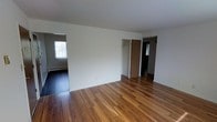 This single-floor residential space offers a clean, functional layout designed for comfortable everyday living. The interior is immediately defined by rich, warm-toned hardwood floors that run throughout, adding character and a sense of continuity across the space.

The main living area is bright and open, with white walls that reflect natural light streaming in from a window in the adjacent room. The neutral palette creates a versatile backdrop, allowing the space to adapt to a variety of furnishing styles and personal tastes.

A built-in closet with a standard door provides convenient storage directly off the main living area, keeping the space organized and clutter-free. A hallway extends further into the unit, leading to additional rooms, including what appears to be a bathroom and a bedroom or secondary space, each accessible through clearly defined doorways.

The adjoining room visible from the main area features darker flooring and a window with a baseboard heating unit below it, suggesting year-round climate comfort. The overall layout flows logically, with rooms connecting in a straightforward manner that maximizes usability.

With its hardwood floors, ample natural light, and well-organized room connections, this space presents a solid and practical living environment ready for occupancy.