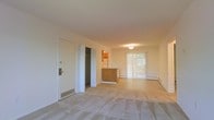 This single-floor space features an open-concept layout that integrates the living area and kitchen into one cohesive space. The living area is finished with light-toned carpeting, contributing to a neutral, understated aesthetic throughout. Walls are painted in a soft, warm white, creating a bright and clean atmosphere.

A peninsula-style kitchen divider with wood-toned cabinetry separates the cooking area from the living space, providing functional counter space while maintaining the open feel of the floor plan. The kitchen area features a refrigerator visible in the background, and the space is accounted for practically within the overall layout.

Near the entry, a closet provides immediate storage, while a baseboard heating unit runs along the far wall, indicating a practical and efficient heating system. A sliding glass door fitted with sheer white curtains allows natural light to filter into the living area, offering a connection to the outdoors. The ceiling features a flush-mount light fixture, complementing the clean, simple design. Overall, the space presents a functional and straightforward layout with a light, neutral palette.