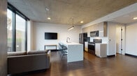 This two-story residence showcases a contemporary industrial aesthetic defined by exposed concrete ceilings, dark hardwood flooring, and a clean, minimalist palette throughout. The main level centers around an open-concept living and kitchen area, where a large marble-topped island with bar seating anchors the space. The kitchen is fully equipped with stainless steel and black appliances, including a gas range, refrigerator, microwave, and dishwasher, all complemented by sleek flat-panel cabinetry. A wall-mounted television and in-unit washer and dryer add to the functionality of the main floor. Natural light filters in through large windows offering urban views, and a striking steel-and-glass staircase connects the two levels, adding an architectural focal point to the space. A half bath is also conveniently located on the main level. Upstairs, two well-proportioned bedrooms each feature hardwood floors, ample closet space, and dedicated study areas. Each bedroom is served by its own full bathroom, both finished with large-format tile walk-in showers enclosed in glass, black hardware accents, and white vanities. The overall ambiance is urban, refined, and functional.