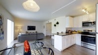 This well-appointed two-bedroom, two-bathroom residence is thoughtfully designed across a single floor, offering a seamless open-concept layout that connects the main living areas. The living room is bright and airy, featuring large windows that invite generous natural light and smooth hardwood-style flooring that extends throughout the space. Clean white walls and a neutral palette create a modern, versatile backdrop, complemented by tasteful décor accents including a large circular mirror and contemporary artwork.

The kitchen is a standout feature, showcasing white shaker-style cabinetry, a dark-topped center island with bar seating, and ample storage. The island serves as both a functional prep area and a casual dining spot, making it ideal for everyday living and entertaining. A dedicated dining area sits adjacent, completing the open social space.

The layout flows naturally between rooms, with wide entryways and consistent flooring reinforcing the cohesive design. The overall ambiance is clean, contemporary, and inviting, with a bright, neutral aesthetic that balances style and practicality throughout this comfortable two-bedroom residence.