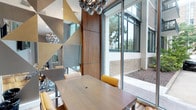 This contemporary single-floor space makes a strong architectural statement through its thoughtful design and high-end finishes. The centerpiece is a sophisticated conference or meeting area anchored by a rich wood-toned table surrounded by sleek chairs with warm mustard yellow upholstery, creating a refined yet inviting atmosphere. Towering floor-to-ceiling glass walls and expansive sliding doors flood the interior with abundant natural light while offering unobstructed views of a well-maintained exterior courtyard, seamlessly blending indoor and outdoor environments. A striking sculptural chandelier with metallic globe clusters serves as a bold focal point overhead, complementing the space's modern aesthetic. Warm walnut wood wall paneling adds depth and texture, contrasting elegantly with the geometric, angular mirror or metallic wall art panels that introduce a dynamic visual element. The overall design language is clean and contemporary, balancing industrial-inspired accents with warm natural materials. The generous ceiling height enhances the sense of openness, making the space feel expansive and airy. This well-appointed environment is ideally suited for professional meetings, collaborative gatherings, or upscale events.