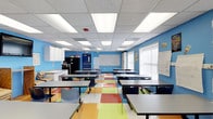 This single-floor educational space is thoughtfully designed to create an energetic and functional learning environment. The room is immediately distinguished by its vivid blue walls, which lend a lively and stimulating atmosphere to the space. Underfoot, a multicolored tile floor in bold shades of red, orange, yellow, and green adds a playful visual element that complements the overall character of the room.

Overhead, a suspended acoustic ceiling grid houses an array of recessed fluorescent panel lights, ensuring bright, even illumination throughout the classroom. Natural light supplements the artificial lighting through large windows fitted with slatted blinds, offering adjustable light control.

Student desks with attached chairs are arranged in organized rows, providing ample seating for a full class. At the front of the room, a large whiteboard serves as the central instructional focal point. Along the side wall, white cabinetry offers storage solutions, accompanied by a utility counter and a refrigerator, adding practical functionality to the space.

Safety features include a clearly marked emergency exit door and a visible fire sprinkler system integrated into the ceiling. Educational displays and materials are posted on the walls, reinforcing the room's dedicated purpose as an active, student-centered learning space.