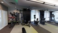 This contemporary fitness center is designed to deliver a complete workout experience within a sleek, modern environment. The space is anchored by a striking large-scale athletic mural that energizes one wall, setting a motivating tone throughout. A dark vertical-panel accent wall provides a bold visual contrast, lending the space a polished, high-end aesthetic.

Natural light fills the room through floor-to-ceiling windows dressed with white roller shades, creating a bright and airy atmosphere that keeps the space feeling open and inviting. Overhead, a ceiling fan and recessed lighting work together to maintain a comfortable environment during workouts.

The fitness center is thoughtfully equipped with a multi-function cable strength training machine, a rowing machine, and multiple cardio units including upright and recumbent stationary bikes, each outfitted with individual monitors. The flooring features a combination of light and dark toned sections, helping to visually define separate workout zones.

The overall ambiance strikes a balance between professional gym functionality and modern interior design, making it an appealing and practical space for a wide range of fitness activities.