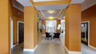 This lobby makes an immediate impression with its vibrant amber-orange walls and crisp white trim, creating a warm and energetic atmosphere. The space is defined by a series of structural columns that frame a central corridor, lending a sense of symmetry and architectural depth. Overhead, tray ceilings with recessed lighting add dimension and a refined touch to the overall design.

The flooring consists of large-format neutral tiles that run throughout, contributing to a clean, polished aesthetic. At the heart of the lobby sits a central seating area anchored by a dark wood table surrounded by sleek black upholstered chairs, arranged atop a textured area rug. This arrangement provides a functional gathering spot while enhancing the lobby's sophisticated character.

Decorative circular metallic wall accents are mounted on the columns on both sides of the corridor, adding an artistic element that complements the bold color palette. Recessed ceiling fixtures provide consistent, bright illumination throughout the space. In the background, glimpses of adjacent areas suggest additional common spaces beyond the main corridor, indicating a well-connected and thoughtfully designed building entrance.