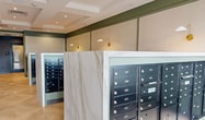 This contemporary mail room, known as the Beck Mail room, is a model of efficient design within its 560 square feet of space. The room boasts a clean, modern aesthetic with a harmonious blend of light and dark elements. Multiple rows of black mailboxes are strategically placed throughout the space, mounted on marble-like partitions that create distinct sections within the room.

The ceiling is a notable feature, with recessed lighting and a coffered design that adds depth and visual interest. The walls are painted in a soothing sage green, complementing the neutral tones of the floor and mailbox units. Large windows and glass doors at one end of the room allow ample natural light to flood the space, enhancing the overall brightness and welcoming atmosphere.

The flooring is particularly striking, featuring a herringbone pattern in light wood tones that adds a touch of sophistication to the utilitarian space. Wall-mounted sconces provide additional lighting and a decorative element. The room's layout is designed for easy navigation, with clear pathways between mailbox sections.

Safety and security features are evident, with visible smoke detectors and likely security cameras integrated into the ceiling design. The overall impression is one of a well-organized, secure, and aesthetically pleasing mail facility that efficiently serves its purpose while maintaining a pleasant environment for users.