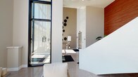 This contemporary building lobby makes an immediate impression with its refined modern design and generous sense of openness. Soaring ceilings amplify the spaciousness of the entry, while an expansive curtain wall of floor-to-ceiling windows framed in dark metal floods the interior with natural light and offers clear views of the surrounding streetscape. The polished light-toned tile flooring reflects the brightness throughout, enhancing the clean, airy aesthetic.

A textured accent wall serves as a focal point, adorned with a cluster of bold, sculptural black floral wall art pieces that add an artistic dimension to the otherwise neutral palette. A classic table lamp with a chrome base completes the vignette, lending a refined touch. White upholstered seating with metallic frames is thoughtfully arranged near the window wall, providing a welcoming area for visitors.

Warm wood-toned wall paneling introduces contrast and warmth to the predominantly white interior, while a sleek elevator bank along the back wall ensures efficient vertical access. The overall ambiance is polished and professional, combining minimalist architecture with carefully selected design details to create a lobby that is both functional and visually striking.