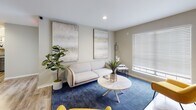 Located at 1200 Halcyon Cir in Greer, South Carolina, this single-floor space is thoughtfully designed to foster comfort, creativity, and community. The main lounge area features warm hardwood-style flooring, multiple seating arrangements, and a lively mix of furnishings including a gray leather armchair, bold yellow striped ottomans, and an olive-green sofa set. Two wall-mounted flat-screen televisions are prominently displayed alongside playful décor, including a "Enjoy the Show" wall script, lending the space an engaging, social atmosphere. A barn-style sliding door adds an architectural accent, while a colorful mural contributes to the energetic personality of the space. A quieter lounge area offers a more refined setting, with a white sofa, blue area rug, abstract canvas artwork, and a round light wood coffee table, complemented by natural light from a window with blinds and lush indoor plants. The kitchen is well-appointed with gray cabinetry, light stone countertops, white subway tile backsplash, open wood shelving, stainless steel appliances, and an industrial-style pendant light. A dedicated restroom is conveniently accessible. Throughout, recessed lighting and neutral wall tones create a clean, welcoming backdrop that ties together the space's eclectic and functional design.