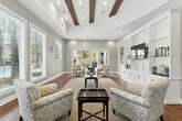 Located at 39415 Ardenwood Way in Lake Elsinore, California, this well-designed single-floor space seamlessly blends elegance and functionality in an open-concept layout. The soaring vaulted ceiling adorned with rich exposed wood beams immediately draws the eye upward, lending a warm architectural character throughout the interior.

The lounge area features warm hardwood flooring, a large woven area rug, and comfortable upholstered seating arranged around a dark wood coffee table. A striking floor-to-ceiling built-in white entertainment unit with glass shelving serves as a focal point along one wall, flanked by framed artwork. A dramatic natural stone accent wall adds texture and visual depth, complementing the neutral gray tones of the walls.

The kitchenette is well-equipped with white cabinetry, dark granite countertops, a double basin sink, and black under-counter appliances. Large windows with white blinds flood the space with abundant natural light, while French doors open directly to an outdoor area, offering views of a pool and patio. Recessed lighting provides additional illumination throughout. The flooring transitions from hardwood in the lounge to clean tile in the kitchen zone, defining each functional area with ease.