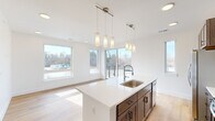 This well-appointed single-floor unit showcases a clean, modern design with an open-concept layout that seamlessly connects the kitchen and living areas. The kitchen is a standout feature, equipped with rich dark-toned cabinetry, stainless steel appliances, a classic white subway tile backsplash, and a spacious center island topped with white quartz countertops. A trio of elegant pendant lights illuminates the island, complemented by recessed ceiling lighting throughout. Large windows and a sliding glass door lead to an outdoor balcony, flooding the living area with abundant natural light and providing views of the surrounding landscape. Light hardwood flooring runs throughout the entire unit, creating a warm and cohesive feel. A well-lit hallway connects the main living space to the private rooms, each finished with the same light hardwood flooring and crisp white walls. The bathroom features a combined tub and shower, a wood-toned vanity with a white countertop, and tile flooring. Additional highlights include a generously sized walk-in closet with built-in shelving and a dedicated laundry area with shelving and access panel. The overall ambiance is bright, airy, and move-in ready.