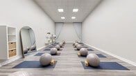 This 790 square foot yoga studio in Indianapolis offers a harmonious blend of functionality and tranquility. The space boasts a modern, minimalist design with a focus on creating a serene atmosphere for practice. Light gray walls are complemented by a stylish wood-look vinyl flooring, creating a neutral backdrop that enhances the room's calming ambiance.

The studio is well-equipped for various yoga practices, featuring neatly arranged blue yoga mats with stability balls and wooden blocks for each participant. Large, arched mirrors line one wall, allowing practitioners to observe and correct their form. Ample storage is provided by white shelving units and wicker baskets, maintaining the room's clutter-free aesthetic.

Soft, flowing curtains frame a feature wall adorned with a minimalist lotus flower design, adding a touch of elegance and reinforcing the space's purpose. The room is illuminated by recessed LED lighting, providing even, glare-free illumination throughout.

The studio's layout is spacious and uncluttered, allowing for easy movement and comfortable spacing between practitioners. A single door provides access, likely leading to additional facilities. This thoughtfully designed space offers an ideal environment for both group classes and individual yoga sessions, promoting focus, balance, and inner peace.