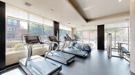 Located at 151 Q St NE in Washington, DC, this fitness center offers a well-appointed exercise facility designed to support a comprehensive workout experience. The space is spread across a single floor and is thoughtfully organized into distinct zones for cardio training, strength work, and functional fitness.

The cardio zone is positioned along a wall of large, floor-to-ceiling windows that flood the area with abundant natural light and provide views of the surrounding neighborhood. A row of treadmills, ellipticals, and stationary bikes lines this sunlit perimeter, creating an open and energizing environment for aerobic exercise.

The strength training area features a robust selection of cable machines, a multi-use functional trainer, and adjustable weight benches, supported by a well-stocked dumbbell rack spanning a wide range of weights. Full-length mirrors line the walls, visually expanding the space while allowing users to monitor their form.

A dedicated functional fitness zone includes medicine balls, resistance bands, and stability equipment, complemented by wall-mounted flat-screen TVs throughout the facility. Cylindrical architectural columns and a tray ceiling add refined structural character to the space. The dark rubberized flooring provides durability and comfort underfoot, while recessed lighting ensures consistent illumination throughout. The overall design balances modern aesthetics with practical, high-performance functionality.