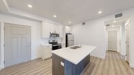 This well-designed 1-bedroom, 1-bathroom unit offers a contemporary layout that maximizes every inch of its single-floor footprint. The open-concept kitchen and living area serves as the heart of the space, anchored by a striking center island with a dark wood-grain base, white quartz countertop, and integrated double sink with a stainless steel dishwasher below. The kitchen features crisp white shaker cabinetry, white quartz countertops, and a full suite of stainless steel appliances including a range, over-the-range microwave, and refrigerator. Light wood-style plank flooring runs throughout the main living areas, creating a clean and cohesive aesthetic. The living area benefits from a sliding glass door with vertical blinds, inviting natural light and providing access to an outdoor space. The bedroom offers plush carpeting, a generous closet, and a large window with blinds that fills the room with natural light. Adjacent to the bedroom, the full bathroom is finished in an all-white palette with a wide vanity, quartz countertop, a large mirror, and a tub-shower combination. Recessed lighting throughout the unit enhances the bright, modern ambiance.