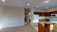 This single-floor residence offers a thoughtfully designed open-concept layout that promotes a natural flow between living and functional spaces. The kitchen is a standout feature, showcasing rich warm-toned wood cabinetry that extends to the ceiling, including a distinctive wine rack detail at the upper level. A full-size refrigerator, light countertops, and ample cabinet storage make the kitchen both practical and inviting. Recessed lighting throughout provides consistent, clean illumination that complements the neutral color palette of soft beige walls and wall-to-wall carpeting in the living area. The open passageway connects the main living space to additional rooms beyond, where natural light pours in through windows, brightening the interior. The layout benefits from a peninsula or breakfast bar area, adding to the kitchen's functionality and providing a casual dining option. The overall ambiance is warm and understated, with a cohesive design language that ties the spaces together through consistent finishes and a light, airy feel. The home's straightforward single-level layout offers convenience and accessibility throughout.