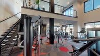 This two-story fitness center in Pearland, TX (77584) is a thoughtfully designed amenity space that combines functionality with contemporary style. The main floor features an expansive workout area outfitted with Matrix-brand cardio equipment, including treadmills, stair climbers, and stationary bikes, arranged to take full advantage of the large floor-to-ceiling windows that frame views of the resort-style pool and surrounding community. The bold geometric-patterned carpet in shades of red, orange, and gray adds a vibrant, energetic aesthetic throughout the strength training zones, which are equipped with cable machines, free weights, and a variety of resistance equipment. Full-length mirrors line key walls, enhancing the sense of openness. A dedicated flex or stretching area features hard surface flooring and exposed industrial ductwork for an urban touch, along with a built-in hydration station set against a striking red-and-gray herringbone accent wall. A sleek metal staircase connects to the second-floor mezzanine level, which overlooks the main workout floor below and offers additional equipment space. The soaring double-height ceilings, recessed lighting, and ceiling fans throughout ensure comfort during any workout. The entrance area includes generous height and glass-panel doors, creating a welcoming and accessible entry experience.