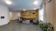 The Mat Morristown presents a thoughtfully designed lobby that serves as an inviting introduction to the space. The focal point of the room is a full-width reclaimed wood accent wall, finished in warm, natural tones with a rustic plank texture that adds character and depth. Prominently displayed on this wall is the branded "M – The Mat Morristown" medallion logo, reinforcing the identity of the space with a polished, professional touch.

The flooring features large-format slate-toned tiles laid in a staggered pattern, providing a durable yet refined surface that complements the earthy warmth of the wood wall. Seating is arranged in a conversational grouping, consisting of a leather sofa in a rich cognac tone, flanked by modern upholstered armchairs in neutral gray. A concrete rectangular coffee table anchors the arrangement, adding an industrial edge to the overall design.

Natural light filters into the space through windows dressed with white blinds, brightening the room alongside recessed and flush-mount ceiling fixtures. Potted greenery is thoughtfully placed throughout, softening the modern aesthetic and adding a welcoming, organic element. The overall ambiance is clean, contemporary, and inviting — striking a balance between professional functionality and comfortable hospitality.