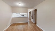 This single-floor residence offers a clean, functional layout designed to maximize everyday livability. The main living area is finished with warm hardwood-style flooring that extends throughout the space, complementing the neutral beige and white tones on the walls and trim. A textured ceiling adds subtle architectural character overhead.

The kitchen is accessible via a pass-through window counter that opens directly into the living area, creating a connected, open feel between the two spaces. White cabinetry lines the kitchen walls, and white appliances — including a refrigerator and range — are neatly integrated into the layout.

Adjacent to the main living space, a hallway leads to additional rooms, with a built-in shelving unit or closet visible along the corridor, offering practical storage solutions. Doorways along the hall provide access to what appear to be a bedroom and additional utility or storage areas.

Natural light filters through the space, keeping the interior bright and welcoming. The overall design is straightforward and well-maintained, with clean lines and a neutral palette that provides a versatile backdrop for a variety of interior styles. The single-level floor plan ensures easy navigation throughout the residence.