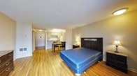This single-floor studio unit offers an efficient, open-plan layout that combines living, sleeping, and cooking functions within one cohesive space. The sleeping area is anchored by a full-sized bed with a dark ornate headboard, accompanied by a bedside table and lamp, creating a functional resting nook. Warm-toned hardwood flooring extends throughout the entire unit, lending a sense of continuity and warmth to the space.

Toward the rear, a compact kitchenette is outfitted with white cabinetry, under-cabinet lighting, and a countertop with a sink, providing essential cooking amenities in a space-conscious design. A small dining set with a table and chairs sits just in front of the kitchen area, offering a dedicated spot for meals.

A hallway near the entrance leads to what appears to be a closet and the unit's entry door, keeping the main living area unobstructed. The neutral gray and beige wall tones complement the flooring, while standard-height ceilings with recessed and under-cabinet lighting maintain a practical, well-lit environment throughout the unit.