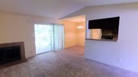 This single-floor residence is designed with an open-concept layout that integrates the living, dining, and kitchen areas into a cohesive and functional living space. The living area is finished with wall-to-wall carpeting in a neutral tone, providing a comfortable and understated foundation throughout. A large sliding glass door dominates one wall of the living space, flooding the interior with natural light and offering views of lush greenery beyond a private balcony. Vertical blinds provide adjustable light control and privacy.

The dining area flows naturally from the living space and is distinguished by a decorative chandelier overhead, adding a touch of warmth and definition to the zone. Neutral-toned walls carry throughout, maintaining a consistent and clean aesthetic. Adjacent to the dining area, the kitchen is positioned behind a raised breakfast bar, creating a partial visual separation while preserving the open feel of the space. A wall-mounted television is pre-installed above the kitchen counter area, adding a practical feature to the layout. The kitchen also includes a stone tile backsplash, contributing a subtle textural contrast to the otherwise smooth finishes of the space.