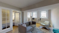 This single-floor residence offers a thoughtfully designed open layout finished in soft neutral tones throughout. The living area serves as the central hub of the space, featuring warm hardwood flooring, a tufted grey sofa, and an accent armchair arranged atop a patterned area rug. Multiple windows allow natural light to fill the room, while varied ceiling heights add architectural interest. A glass-paneled French door connects the living area to the private bedroom, which features a tray ceiling with integrated lighting, hardwood floors, and ample room for a queen-sized bed with an upholstered headboard. The kitchen is efficiently designed with white cabinetry, stone countertops, a grey tile backsplash, stainless steel appliances including a built-in oven and microwave, and a large window framing an outdoor view. A bar-height counter with seating provides a casual dining option adjacent to the kitchen. The bathroom is finished in a clean, modern style with large-format tile flooring, a floating vanity, a wide mirror with overhead lighting, and a full sofa tub with shower. A hallway connects the rooms, completing the functional and cohesive floor plan.