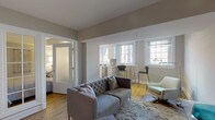 This single-floor residence offers a thoughtfully designed open layout finished in soft neutral tones throughout. The living area serves as the central hub of the space, featuring warm hardwood flooring, a tufted grey sofa, and an accent armchair arranged atop a patterned area rug. Multiple windows allow natural light to fill the room, while varied ceiling heights add architectural interest. A glass-paneled French door connects the living area to the private bedroom, which features a tray ceiling with integrated lighting, hardwood floors, and ample room for a queen-sized bed with an upholstered headboard. The kitchen is efficiently designed with white cabinetry, stone countertops, a grey tile backsplash, stainless steel appliances including a built-in oven and microwave, and a large window framing an outdoor view. A bar-height counter with seating provides a casual dining option adjacent to the kitchen. The bathroom is finished in a clean, modern style with large-format tile flooring, a floating vanity, a wide mirror with overhead lighting, and a full sofa tub with shower. A hallway connects the rooms, completing the functional and cohesive floor plan.