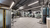 Located at 118 Park Ave, this fitness center offers a comprehensive workout environment designed with both functionality and aesthetic appeal in mind. The space features an industrial-modern design, with an exposed ceiling showcasing dark-painted ductwork and structural elements alongside rows of linear LED lighting that illuminate the floor evenly and effectively.

The main workout area is generously sized, providing ample room to move between stations. Durable rubber tile flooring spans the entire space, offering comfort and safety underfoot. The strength training zone is well-appointed with a multi-functional cable machine, a Smith machine-style arch rack with an adjustable bench, a full dumbbell rack, kettlebells, medicine balls, TRX suspension trainers, and a heavy punching bag. Full-length mirrors along multiple walls enhance the sense of space and allow for form monitoring during exercise.

The cardio section is lined along a striking illustrated mural featuring athletic figures in green and black tones, adding visual energy to the room. Treadmills, ellipticals, and rowing machines are arranged in front of mounted flat-screen televisions. A glass-walled enclosure near the entrance provides a defined transition zone, and a water station is conveniently accessible. The overall ambiance is dynamic, focused, and professionally appointed.