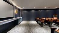 Located at 1800 Palmer Rd in Fort Washington, MD, this dedicated screening room offers a refined, cinema-inspired environment designed for an immersive viewing experience. The room is finished in deep navy blue walls that create an intimate, theater-like atmosphere, complemented by recessed ceiling lighting that provides even, ambient illumination throughout the space. The flooring consists of a textured, multi-toned carpet that adds acoustic warmth while reinforcing the room's polished aesthetic. Rows of theater-style seats with rich cognac leather upholstery and fold-down armrests are arranged to face a large projection screen mounted on the front wall, which is framed by decorative white molding panels and accented by striking gold wall sconces. A built-in low-profile cabinet runs along the front wall beneath the screen, offering practical storage while maintaining a clean, streamlined look. A standout feature is the classic red popcorn machine stationed at the back of the room, adding a fun, authentic touch to the cinematic setting. The space is accessed through a solid paneled door and is equipped with a standard HVAC system and emergency exit signage, ensuring both comfort and safety. This well-appointed screening room delivers a compact yet fully realized private theater experience.