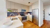 Located at 3025 John F Kennedy Blvd in Philadelphia's University City neighborhood, this modern high-rise residence offers a thoughtfully designed single-floor layout with clean contemporary aesthetics throughout. A welcoming entry hallway with warm hardwood flooring and crisp white walls leads into the main living areas, offering a first glimpse of the sweeping city skyline beyond.

The living room is a standout space, anchored by expansive floor-to-ceiling windows that frame breathtaking panoramic views of the Philadelphia skyline. Natural light floods the room, complementing the light hardwood floors and white walls adorned with bold abstract artwork. The space is well-proportioned and flows seamlessly from the living area into the kitchen.

The kitchen is outfitted with flat-front cabinetry in a warm wood tone, white quartz countertops, a decorative tile backsplash, and a full suite of stainless steel appliances, combining both style and functionality. The bedroom continues the residence's light-filled theme, featuring its own floor-to-ceiling window with striking city views, hardwood flooring, and a serene, minimalist atmosphere. A secondary interior bedroom provides a quiet, restful retreat. The bathroom is finished with neutral tile, a wood-toned vanity, a tall framed mirror, and warm sconce lighting, achieving a clean and modern look.