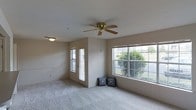 This single-floor residential space offers a welcoming and functional layout designed for comfortable everyday living. The main living area is generously proportioned, featuring soft wall-to-wall carpeting that adds warmth underfoot and contributes to the room's calm, neutral aesthetic. The space is bathed in natural light, thanks to multiple large windows, including a wide multi-pane window that frames a pleasant outdoor view of lush greenery and mature trees. A glass-panel exterior door further enhances the connection to the outdoors while allowing additional light to fill the room. The walls are finished in a soft, neutral tone that complements the overall airy feel of the space. A brass ceiling fan with wooden blades is centrally positioned overhead, providing both practical airflow and a classic decorative touch, while a secondary light fixture near the entry ensures the space remains well-lit throughout. The overall ambiance is light, open, and understated, making it a versatile canvas well-suited to a variety of interior styles. The single-floor layout promotes ease of movement and accessibility throughout the home, creating a cohesive and connected living environment.