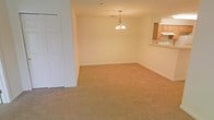This single-floor space features an open-concept layout that seamlessly connects the living, dining, and kitchen areas. The main living and dining area is finished with wall-to-wall beige carpeting, lending warmth and comfort underfoot. Neutral cream-toned walls keep the space feeling bright and versatile, offering a clean backdrop adaptable to a variety of interior styles.

A pendant chandelier with a classic multi-bulb design hangs centrally over the dining area, serving as a focal point and providing ambient overhead lighting. The kitchen is accessible via a raised passthrough counter, which visually connects the cooking space to the main living area while maintaining a degree of separation. Honey-toned wood cabinetry is visible from the passthrough, contributing a warm, traditional aesthetic to the kitchen. A white door with standard paneling is positioned along one wall, providing access to an additional room or closet.

Overall, the space presents a straightforward, functional layout with a cohesive neutral palette. The open flow between the living, dining, and kitchen zones makes it well-suited for everyday living and casual entertaining.