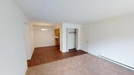This single-floor unit offers an open and functional layout that connects the main living area to the dining space and kitchen. The living room is finished with wall-to-wall carpeting, providing a warm and comfortable feel underfoot, while the transition into the dining and kitchen areas features dark hardwood-style flooring that adds contrast and visual interest throughout the space.

An open closet situated between the living and entry areas offers convenient built-in storage, while a standard entry door with brass hardware provides access to the unit. The dining area is illuminated by a classic chandelier, adding a modest decorative touch to the space.

The kitchen is partially visible from the main living area and features warm wood cabinetry, a stainless steel refrigerator, and a dark countertop. The open sightlines between the kitchen and living spaces contribute to an airy, connected feel throughout the unit.

White walls and a textured white ceiling are consistent throughout, maintaining a clean and neutral palette. A baseboard heating unit along the wall ensures year-round comfort. Overall, the space is straightforward and practical in its design.