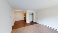 This single-floor unit offers an open and functional layout that connects the main living area to the dining space and kitchen. The living room is finished with wall-to-wall carpeting, providing a warm and comfortable feel underfoot, while the transition into the dining and kitchen areas features dark hardwood-style flooring that adds contrast and visual interest throughout the space.

An open closet situated between the living and entry areas offers convenient built-in storage, while a standard entry door with brass hardware provides access to the unit. The dining area is illuminated by a classic chandelier, adding a modest decorative touch to the space.

The kitchen is partially visible from the main living area and features warm wood cabinetry, a stainless steel refrigerator, and a dark countertop. The open sightlines between the kitchen and living spaces contribute to an airy, connected feel throughout the unit.

White walls and a textured white ceiling are consistent throughout, maintaining a clean and neutral palette. A baseboard heating unit along the wall ensures year-round comfort. Overall, the space is straightforward and practical in its design.