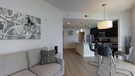 This contemporary single-floor residence offers a clean, open-concept layout designed for comfortable everyday living. The living area features light gray upholstered seating, light wood-tone flooring, and crisp white walls accented by modern wall art, creating a bright and welcoming atmosphere throughout. Recessed lighting illuminates the space seamlessly, complemented by pendant lights that add a refined touch over the kitchen area. The kitchen is well-appointed with dark cabinetry, stainless steel appliances including a refrigerator and microwave, and a light-toned countertop island that doubles as a breakfast bar with sleek black bar stools. A pendant drum light above the dining area adds warmth and definition to the space. The open sightlines between the living and kitchen areas enhance the sense of spaciousness, making the layout ideal for both relaxed daily use and casual entertaining. Light wood-look flooring flows consistently throughout, tying the spaces together cohesively. The neutral color palette of whites, grays, and dark accents gives the residence a modern, polished aesthetic while maintaining a functional and inviting character.