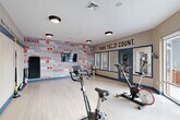 Located at 9142 Richmond Hwy in Fort Belvoir, Virginia, this fitness room is a purpose-built exercise space designed to inspire and energize its users. The room features light hardwood-style flooring throughout, providing a clean and functional surface well-suited for a variety of workout activities.

The space is outfitted with multiple stationary cycling bikes and a weight bench, catering to both cardio and strength training. Stability balls of varying sizes are neatly stored on a dedicated rack, while TRX suspension trainers are mounted directly to the wall, offering versatile bodyweight exercise options. A wall-mounted television is also present, enhancing the workout experience.

The design is defined by bold, motivational typography displayed prominently along the walls. A vivid graphic accent wall features words such as "Inspire," "Engage," "Performance," and "Connect," while a contrasting beige wall carries the phrase "Make Today Count." A continuous motivational quote runs as a border stripe along the upper portion of one wall, reinforcing an energetic atmosphere.

Large sliding glass doors along one wall flood the space with natural light and offer views of the surrounding exterior. Flush-mount ceiling lights provide consistent overhead illumination. The overall ambiance is dynamic, focused, and welcoming — a well-considered fitness environment suited for daily use.
