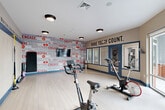Located at 9142 Richmond Hwy in Fort Belvoir, Virginia, this fitness room is a purpose-built exercise space designed to inspire and energize its users. The room features light hardwood-style flooring throughout, providing a clean and functional surface well-suited for a variety of workout activities.

The space is outfitted with multiple stationary cycling bikes and a weight bench, catering to both cardio and strength training. Stability balls of varying sizes are neatly stored on a dedicated rack, while TRX suspension trainers are mounted directly to the wall, offering versatile bodyweight exercise options. A wall-mounted television is also present, enhancing the workout experience.

The design is defined by bold, motivational typography displayed prominently along the walls. A vivid graphic accent wall features words such as "Inspire," "Engage," "Performance," and "Connect," while a contrasting beige wall carries the phrase "Make Today Count." A continuous motivational quote runs as a border stripe along the upper portion of one wall, reinforcing an energetic atmosphere.

Large sliding glass doors along one wall flood the space with natural light and offer views of the surrounding exterior. Flush-mount ceiling lights provide consistent overhead illumination. The overall ambiance is dynamic, focused, and welcoming — a well-considered fitness environment suited for daily use.