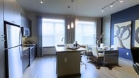This single-floor space showcases a thoughtfully designed open-concept layout that seamlessly integrates the kitchen and dining areas. The kitchen is anchored by a central island equipped with an undermount sink, dark countertops, and bar seating, making it a functional hub for both cooking and casual gatherings. Sleek cabinetry and stainless steel appliances line the kitchen perimeter, contributing to the space's modern aesthetic.

Overhead, a trio of elegant glass pendant lights hangs above the island, complementing the track lighting installed along the ceiling. The warm-toned hardwood flooring runs throughout, adding a natural contrast to the contemporary grey and neutral color palette of the walls and cabinetry.

Large windows fitted with horizontal blinds and floor-length curtains bathe the interior in abundant natural light, enhancing the open and airy feel of the space. The adjacent dining area accommodates a full dining table and chairs, set atop a textured area rug that defines the zone. Overall, the space balances stylish design with practical functionality.