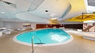 Located at 100 Cold Spring Rd in Rocky Hill, Connecticut, this expansive single-floor aquatic facility spans an impressive footprint designed to accommodate both active and leisure use. The centerpiece is a large, oval-shaped indoor swimming pool with vivid turquoise water, bordered by a broad pool deck finished in textured aggregate flooring. Adjacent to the main pool, a separate hot tub provides a dedicated space for relaxation.

The architecture features a dramatic curved ceiling above the pool area, accented by skylights that introduce natural light and create an open, airy atmosphere. Warm wood slat wall paneling lines the perimeter, adding a resort-like character that complements the crisp white lounge furnishings, including daybeds, chaise lounges, and rounded ottomans with nautical navy-and-white striped accents.

A glazed solarium alcove connects the interior to views of the surrounding greenery, with floor-to-ceiling windows and a translucent roof structure bathing the lounge area in natural light. A separate viewing or relaxation room, separated by large glass panels, offers additional seating with direct sightlines to the pool. Yellow cantilever umbrellas add a vibrant, resort-inspired touch throughout the space. Entry is provided via glass double doors, and the overall ambiance is bright, modern, and welcoming.