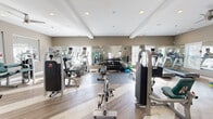 Located at 71 Mayfair Square in Danbury, Connecticut, this fitness center offers a spacious, well-appointed exercise room designed to support a full range of workout routines. The room features light hardwood-style flooring, neutral taupe walls, and a tray ceiling with recessed lighting complemented by a ceiling fan, creating a clean and energizing atmosphere. Large windows along multiple walls allow abundant natural light to fill the space, while full-length mirrors strategically placed throughout enhance the sense of openness and allow users to monitor their form during workouts. The cardio section is equipped with multiple treadmills, elliptical machines, and a stationary bike, catering to a variety of aerobic training preferences. The strength training area includes a comprehensive dumbbell rack with a wide range of weights, adjustable benches, and several cable and resistance machines with teal upholstered seating. Stability balls and balance training accessories are also available, rounding out the functional fitness offerings. The floor plan flows smoothly across the single level, with dedicated mat areas for stretching and floor exercises. A wall-mounted television adds a convenient entertainment option. This thoughtfully equipped fitness center provides everything needed for a complete and effective workout experience.